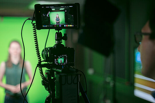 Eine Kamera die eine Person des Medienlabors vor einem Greenscreen filmt.