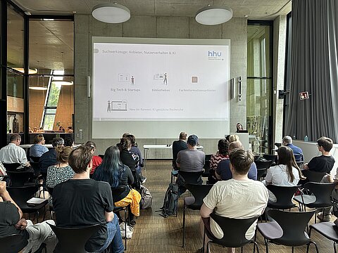 Das Publikum beim E-Learning-Netzwerktreffen in der OASE von hinten, vorne ist eine Präsentation auf dem Beamer zu sehen und am Stehpult eine Rednerin. 