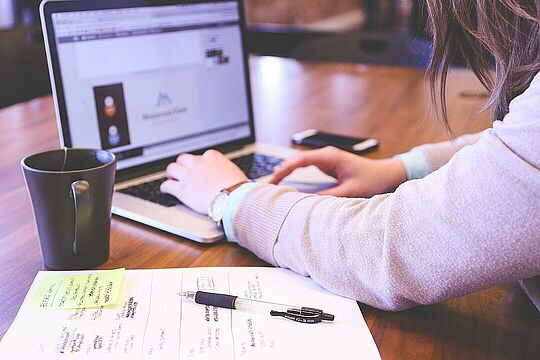 Bild einer Person am Schreibtisch. Sie arbeitet an einem Laptop, um sie herum liegen ein Handy, Unterlagen mit Notizen und ein USB-Stick und neben dem Laptop steht eine Tasse.