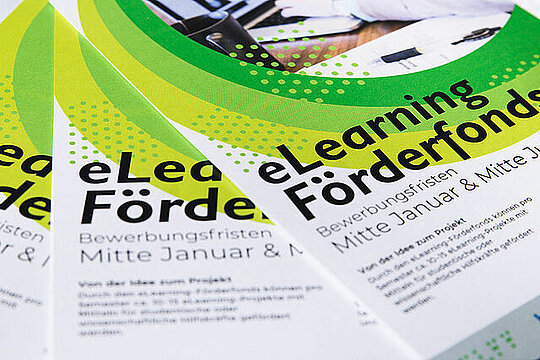 Nahaufnahme von einem aufgefächerten Stapel Flyer mit der Aufschrift "e-Learning Förderfonds".  Darunter in kleinerer Schrift: "Bewerbungsfristen Mitte Januar & Mitte Juni".